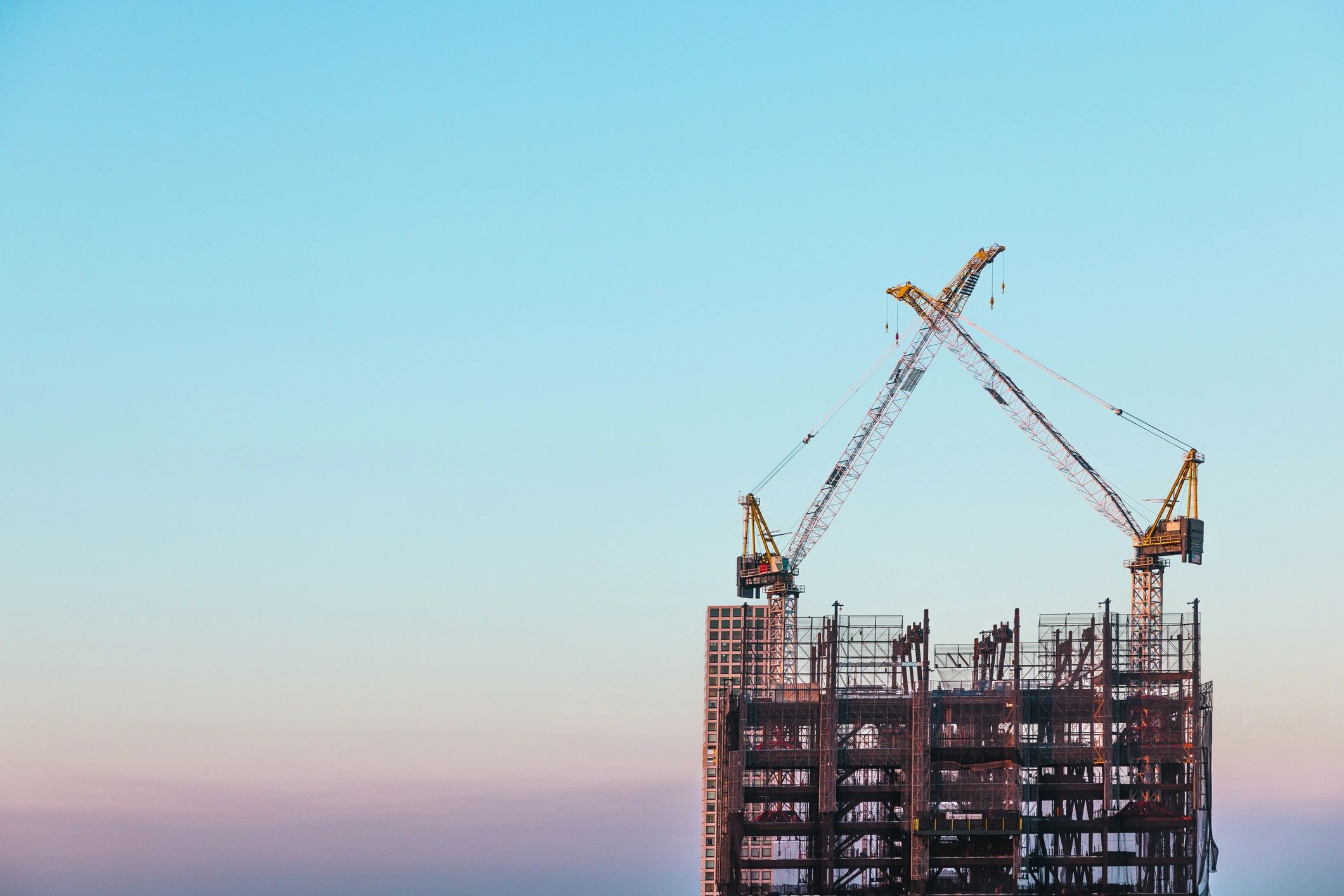 A commercial building under construction against a dusk sky, cranes overhead